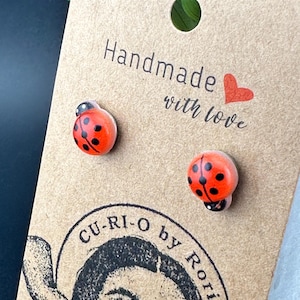 May include: A pair of ladybug stud earrings on a brown card with the text "Handmade with love". The earrings are red with black spots and black heads. The card also has a logo that says "CU-RI-O by Rori".