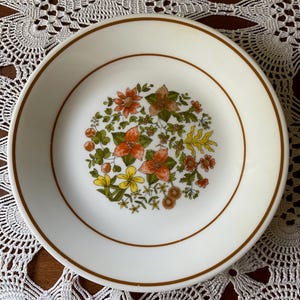 May include: A white ceramic plate with a brown rim and a floral design in the center. The flowers are orange, yellow, and brown, with green leaves. The plate is on a white lace tablecloth.