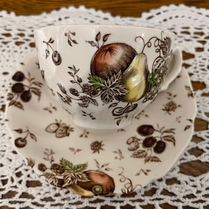 May include: A white teacup and saucer set with a fruit design. The cup and saucer feature brown illustrations of apples, pears, grapes, and berries. The set is placed on a white lace tablecloth.