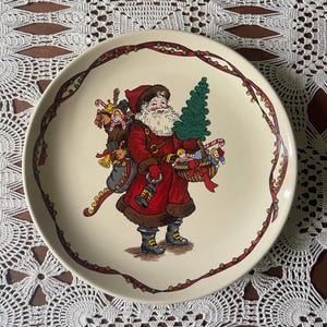 May include: Cream-colored ceramic plate featuring a festive Santa Claus illustration. Santa is depicted in a red coat, carrying a sack of toys and a basket with a Christmas tree. The plate has a red ribbon border with gold accents.