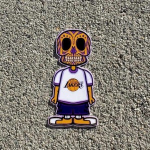 May include: A yellow and purple cartoon skull wearing a white t-shirt with the Los Angeles Lakers logo and a purple basketball. The skull is wearing purple shorts and yellow shoes.