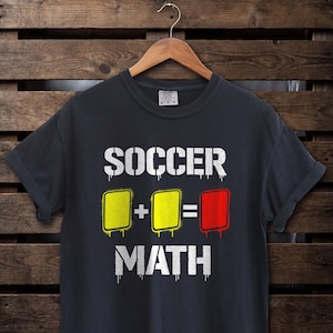 Może przedstawiać: Ciemnoszara koszulka z napisem "SOCCER MATH" w kolorze białym, z kroplami. Projekt zawiera grafikę żółtych i czerwonych kartek, reprezentującą motyw piłkarski. Koszulka jest prezentowana na drewnianym wieszaku.