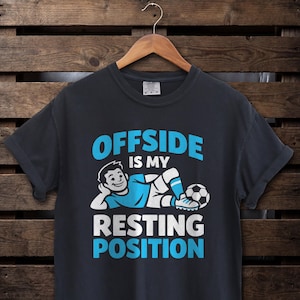 Op de afbeelding: Donkergrijs t-shirt met de tekst "OFFSIDE IS MY RESTING POSITION" in blauw en wit. Een cartoonfiguur in voetbaltenue ligt naast een voetbal. Het t-shirt hangt aan een houten hanger.