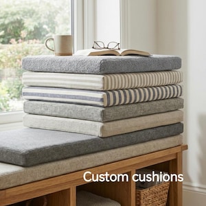 以下が含まれることがあります： さまざまなグレーとベージュの色合いのカスタムクッションのスタック。ストライプと無地のパターンがあります。マグカップ、メガネ、開いた本が上にあります。クッションは窓の近くの木製のベンチに展示されています。