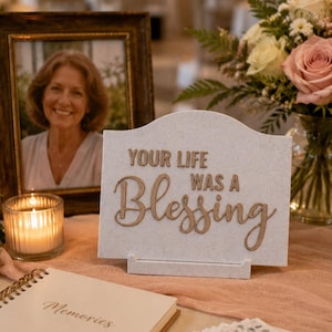 May include: A white speckled rectangular sign with a curved top edge featuring the raised brown text "YOUR LIFE WAS A Blessing" centered on the front. The sign is supported by a small white stand and is positioned on a light pink fabric surface.