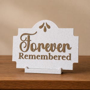 May include: A white plaque with a speckled texture featuring the brown text Forever Remembered in a mix of script and serif fonts. The plaque has a decorative arched top with three brown teardrop shapes and rests on a matching white stand.