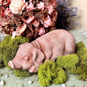 May include: A small, light pink pig figurine lying down, nestled among bright green moss. Behind the pig sits a spherical ornament composed of dried pink flowers, brown botanical elements, and a single pale pink flower, with purple sprigs.