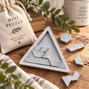 May include: A triangular concrete puzzle frame containing several gray geometric puzzle pieces, accompanied by a beige drawstring fabric pouch printed with the text MINI PUZZLE, SMALL PUZZLES BIG MINDS, and a small heart icon, with extra pieces nearby.