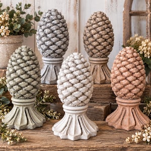 May include: A collection of five decorative pinecone sculptures, each featuring a textured, layered scale pattern and mounted on a fluted pedestal base. The sculptures are finished in various muted tones including grey, beige, white, green, and terracotta.