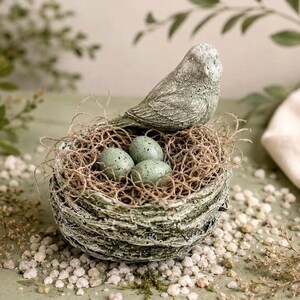 May include: A decorative bird figurine with a weathered green and gray finish perched on the edge of a textured, bowl-shaped nest. The nest contains three speckled, light green eggs nestled within a bed of thin, brown, fibrous material.