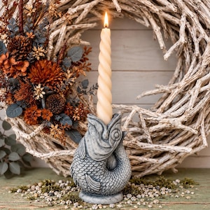 May include: A gray stone-textured fish-shaped candle holder with a spiral-patterned cream-colored taper candle. The fish is depicted with detailed scales and an open mouth, serving as the base for the lit candle, creating a vertical focal point.