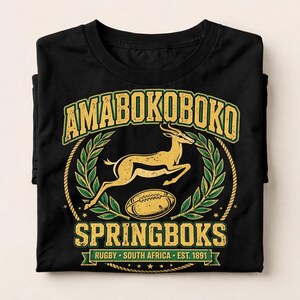 Op de afbeelding: Een zwart t-shirt met een geel en groen grafisch ontwerp van een springende springbok en een rugbybal. De tekst luidt AMABOKO BOKO, SPRINGBOKS en RUGBY SOUTH AFRICA EST. 1891. Het ontwerp is omkaderd door een lauwerkrans en touw.