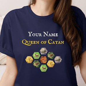 May include: Navy blue t-shirt with the text "Your Name Queen of Catan" in white and yellow. The shirt features a graphic of hexagonal shapes with resource icons. A casual wear item.