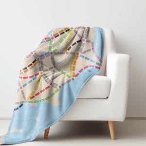 May include: A blue and beige throw blanket with a colorful subway map design. The blanket is draped over a white armchair with wooden legs.