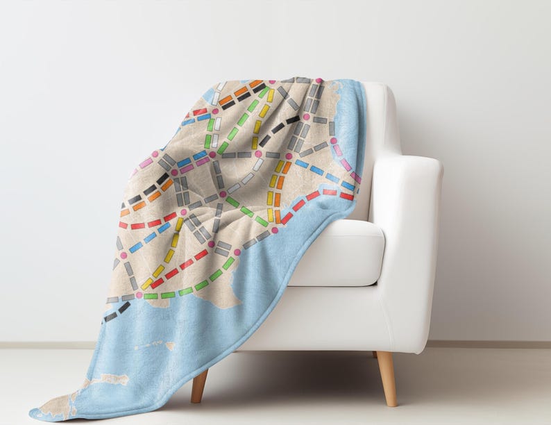May include: A blue and beige throw blanket with a colorful subway map design. The blanket is draped over a white armchair with wooden legs.