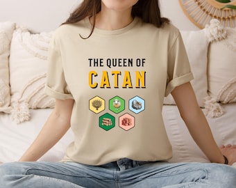 May include: A beige t-shirt with the text "The Queen of Catan" in yellow letters. The shirt features a graphic of six hexagonal tiles representing the resources in the board game Catan.