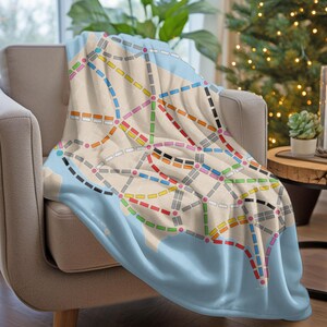 May include: A blue and white throw blanket with a colorful subway map design. The blanket is draped over a light brown armchair.