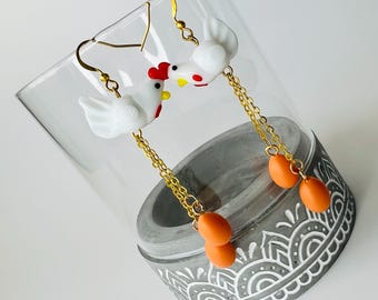 Pode incluir: Um par de brincos pendurados e extravagantes com figuras de galinha brancas com cristas vermelhas e bicos amarelos. Os brincos têm correntes douradas e pingentes em forma de ovo laranja. São exibidos em um copo transparente.