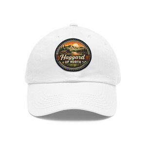 May include: A white baseball cap featuring a circular patch on the front. The patch displays a landscape illustration of mountains, a lake, and a cabin at sunset, with the text "Haggard UP NORTH" and two small crossed axes printed below.