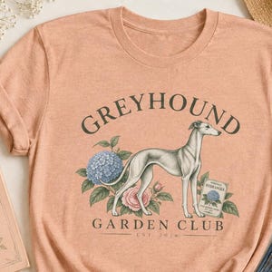 以下が含まれることがあります： 花の中に立つグレーハウンドのグラフィックが描かれたピーチカラーのTシャツ。テキストには「GREYHOUND GARDEN CLUB EST 2024」と書かれています。デザインには青いアジサイ、ピンクのバラ、青い花が描かれた小さな長方形の看板が含まれています。