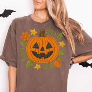 May include: Brown t-shirt featuring an embroidered orange jack-o'-lantern with a smiling face, surrounded by green vines, yellow flowers, and a small pumpkin. The shirt is a relaxed fit, perfect for Halloween.