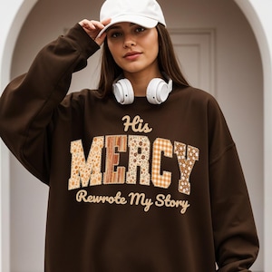 Puede incluir: Una sudadera marrón con el texto "His MERCY Rewrote My Story" en un diseño de patchwork. La sudadera se usa con una gorra de béisbol blanca y auriculares.