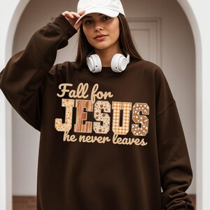 Op de afbeelding: Een donkerbruine sweatshirt met de tekst "Fall for JESUS he never leaves". De tekst is in een mix van lettertypen en patronen. De sweatshirt heeft een ronde hals en lange mouwen.