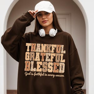 以下が含まれることがあります： パッチワークデザインで「THANKFUL, GRATEFUL, BLESSED」の文字が入った茶色のスウェットシャツ。「God is faithful in every season」の文字が下にあります。白い野球帽とヘッドホンを着用しています。