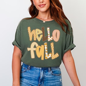 以下が含まれることがあります： オリーブグリーンのTシャツ。"hello fall"の文字がパッチワークデザインで描かれています。文字は黄色、オレンジ、茶色など、さまざまな秋の色と模様で構成されています。カジュアルな半袖スタイルです。
