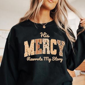 Puede incluir: Sudadera negra con el texto "His MERCY Rewrote My Story". La palabra "MERCY" está en un diseño de patchwork con estampados florales y a cuadros. La sudadera es de estilo casual.