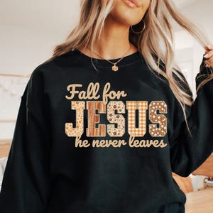 以下が含まれることがあります： 「Fall for JESUS he never leaves」の文字が入った黒いスウェットシャツ。「JESUS」の文字は、花柄、チェック柄、木目模様のパッチワークデザイン。秋をテーマにした宗教的なアパレルアイテム。