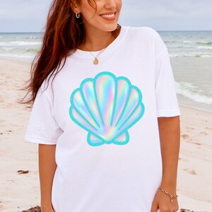 May include: White t-shirt featuring a large, iridescent seashell graphic with a turquoise outline. The seashell design has a shimmering, holographic effect. The shirt is a casual, oversized style, perfect for beach or summer wear.