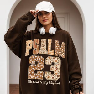 May include: A brown sweatshirt with the text "PSALM 23" in a floral and plaid design. The number "23" is also in a floral pattern. The phrase "The Lord Is My Shepherd" is printed below the numbers.
