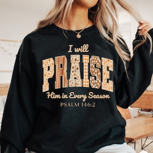 Puede incluir: Sudadera negra con la frase "I will PRAISE Him in Every Season". La palabra "PRAISE" tiene un diseño de patchwork con estampados florales y a cuadros. El texto "PSALM 146:2" está impreso debajo.