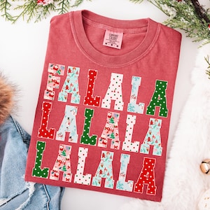 Può includere: Una maglietta rossa Comfort Colors con la scritta "FALALA" in un motivo ripetuto. Le lettere sono rosse, verdi e bianche con motivi festivi. La maglietta è piegata su una superficie bianca con decorazioni natalizie.