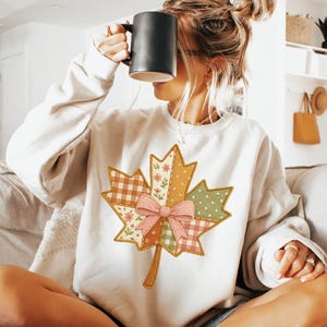 Könnte beinhalten: Cremefarbenes Sweatshirt mit einem großen Ahornblatt-Design im Patchwork-Stil und einer rosa Schleife. Das Blatt enthält verschiedene Muster, darunter Blumen, Vichy-Karo und Punkte. Das Sweatshirt hat lange Ärmel und einen Rundhalsausschnitt.