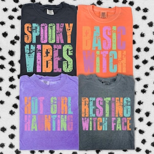 Könnte beinhalten: Vier gefaltete T-Shirts in verschiedenen Farben, jedes mit einem anderen Satz in farbenfrohen, verwitterten Buchstaben. Die Sätze lauten "Spooky Vibes", "Basic Witch", "Hot Girl Haunting" und "Resting Witch Face". Die Shirts befinden sich auf einem schwarz-weiß gepunkteten Hintergrund.