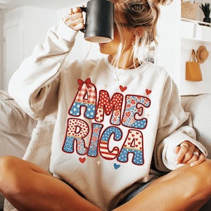 May include: Cream-colored sweatshirt with the word "AMERICA" in a colorful, patriotic design. The letters feature patterns like gingham, stars, and floral prints. A black mug is held up to the face.