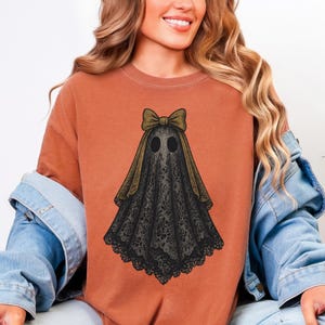 K&ouml;nnte beinhalten: Rostfarbenes T-Shirt mit einem schwarzen und goldenen Geisterdesign. Der Geist hat ein spitzenartiges Muster und eine Schleife auf dem Kopf. Das Design ist auf dem T-Shirt zentriert und schafft eine Halloween-&Auml;sthetik.