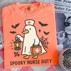 May include: Orange t-shirt with a cartoon ghost duck wearing a nurse's hat and holding a lantern and medical bag. The shirt has the text "SPOOKY NURSE DUTY" and is decorated with bats, stars, and a spiderweb. The shirt is a Comfort Colors brand.