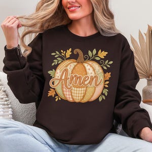 May include: A brown sweatshirt featuring a pumpkin design with the word "Amen" in a decorative font. The pumpkin is composed of various patterns and colors, surrounded by autumn leaves. The sweatshirt is a fall-themed apparel item.