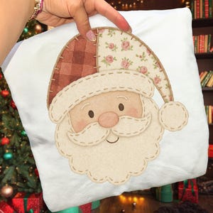 May include: White square pillow featuring a Santa Claus design. The Santa has a beige beard and rosy cheeks, with a hat made of patchwork fabric in a floral pattern and a plaid design. The pillow is held by a person.