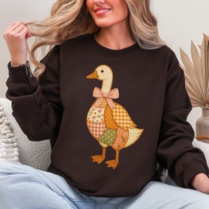 May include: A dark brown sweatshirt featuring a whimsical goose design. The goose is adorned with a patchwork of floral, gingham, and dotted patterns in shades of orange, green, and cream. A pink bow accents the goose's neck, adding a touch of charm to this cozy apparel.