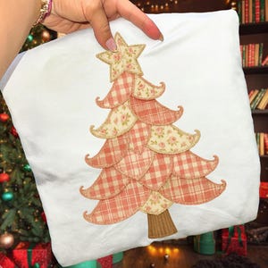 May include: White square pillow with a Christmas tree design. The tree is made of alternating layers of peach gingham and floral fabric, topped with a star. The tree trunk is brown.