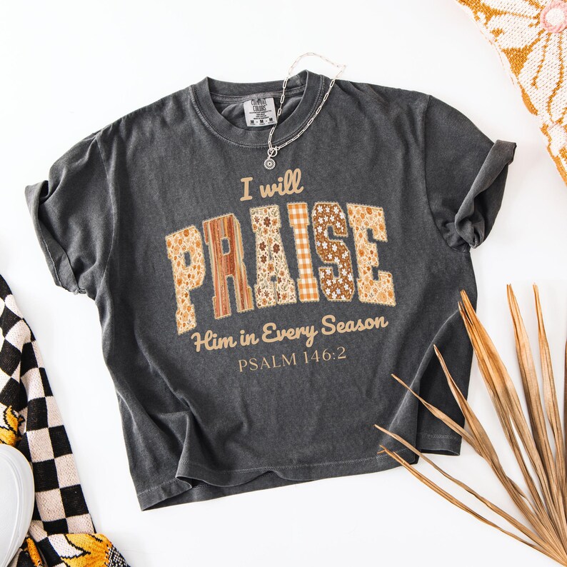 May include: Dark gray t-shirt with the words "I will PRAISE Him in Every Season" in a patchwork design. The word "PRAISE" is in a mix of floral and plaid patterns. The text "PSALM 146:2" is below the main text.