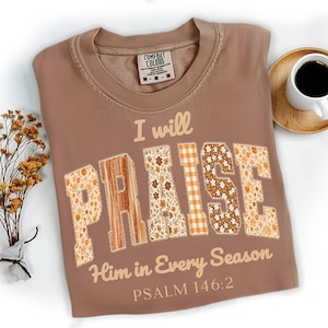 May include: A brown Comfort Colors t-shirt with the words "I will PRAISE Him in Every Season" and Psalm 146:2 printed on it. The word "PRAISE" is in a patchwork design with floral and plaid patterns. A cup of coffee sits on a wooden coaster.