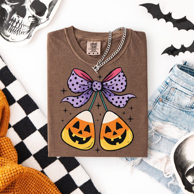 May include: A brown Comfort Colors t-shirt featuring a Halloween-themed graphic. The design includes a purple bow with polka dots, two candy corn pieces with jack-o'-lantern faces, and small star accents. A silver chain necklace is draped across the shirt.