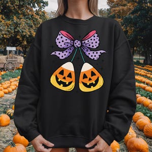 May include: Black sweatshirt featuring a Halloween-themed graphic. The design includes two candy corn pieces with jack-o'-lantern faces, connected by a purple bow with pink and white polka dots. The background shows a pumpkin patch.