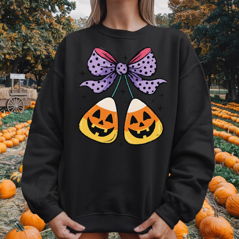 May include: Black sweatshirt featuring a Halloween-themed graphic. The design includes two candy corn pieces with jack-o'-lantern faces, connected by a purple bow with pink and white polka dots. The background shows a pumpkin patch.