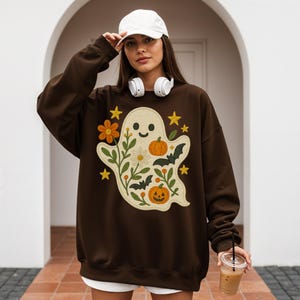 May include: A dark brown sweatshirt with a playful ghost design, including pumpkins, flowers, bats, and stars. The ghost has a smiling face and is surrounded by floral elements. The sweatshirt is a comfortable, casual style.